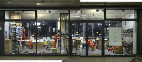 Nachtansicht einer gläsernen Werkstatt, Frau in Orange steht auf einem Hocker und reckt sich, um an ein hohes Regalfach zu gelangen