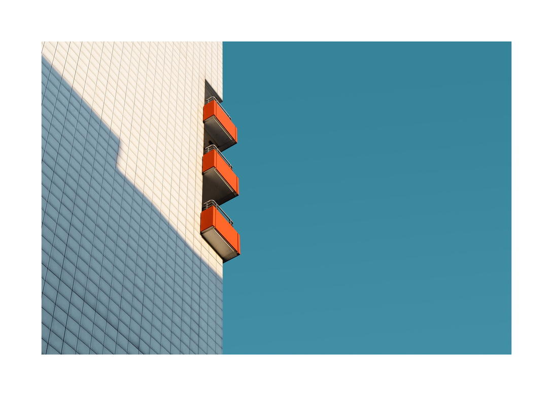 drei rot verkleidete Balkone an einem weiß gekachelten Hochhaus vor flächig blauem Himmel