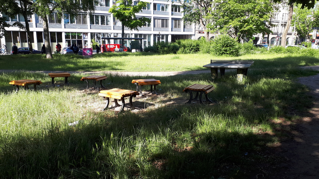 Grünfläche mit Tischtennisplatte und Sitzhockern, im Hintergrund auf der anderen Straßenseite das Aufbau Haus