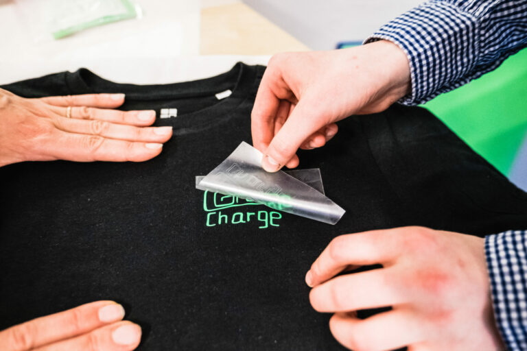 Hände bringen mittels einer Folie einen grünen Schriftzug auf ein schwarzes T-Shirt auf