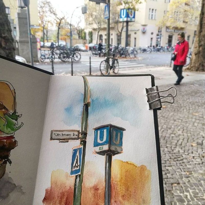 Skizzenbuch aufgeschlagen mit Aquarell Zeichnung von U-Bahn- und Straßenschild, im Hintergrund die reale Szenerie
