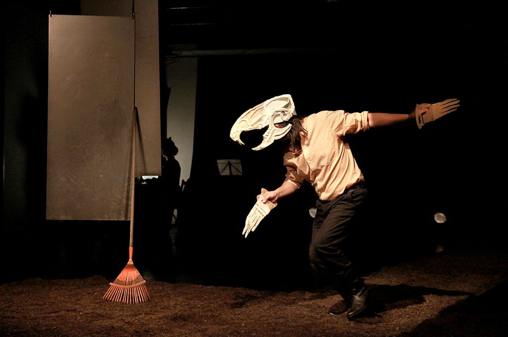 Bühnensituation, Mensch mit der Maske eines Tierschädels und aus Pappe gebastelten Pfoten in  Bewegung auf einem mit Stroh ausgelegten Boden