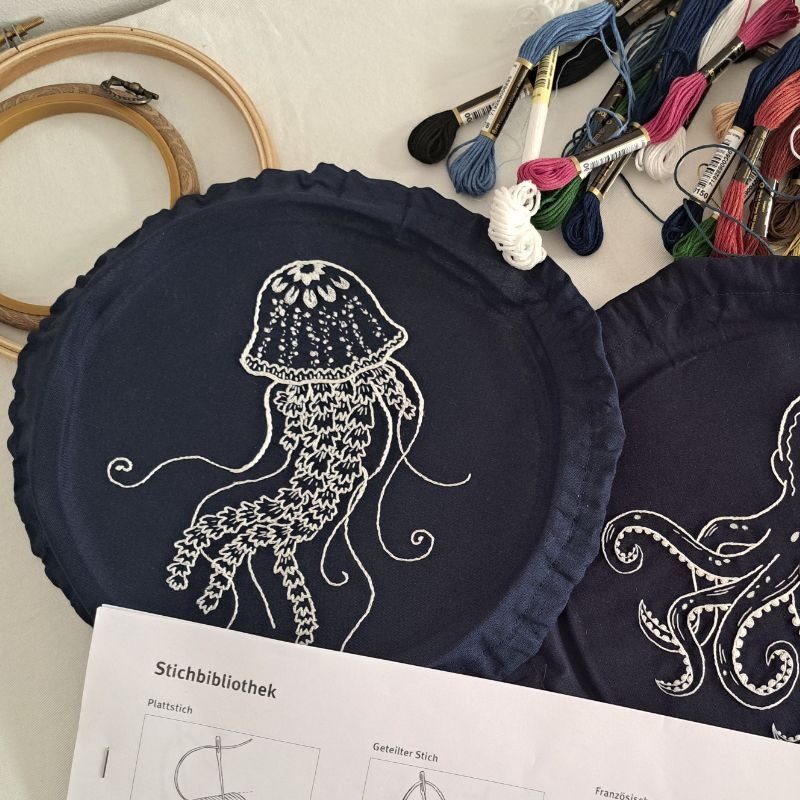 Foto einer Stickarbeit, dunkelblaue Fläche mit weißer Qualle, daneben liegt verschiedenfarbiges Garn auf einem hellen Untergrund