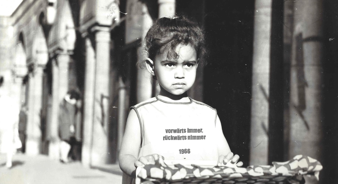 schwarz weiß Foto eines kleinen Mädchens mit zusammengebundenen, dunklen Haaren, das direkt in die  Kamera schaut. sie trägt ein Shirt auf dem steht: Vorwärts immer, rückwärts nimmer 1966