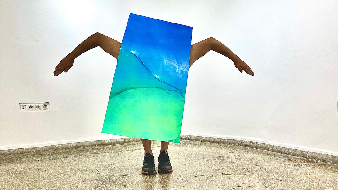 Mensch in einer performativen Bewegung hinter einem Bild aus blau-grünem Fabrverlauf, in der Ecke eines Raumes mit hellem Steinboden und weißen Wänden. von dem Menschen sind nur Arme und Füße in Schuhen sichtbar