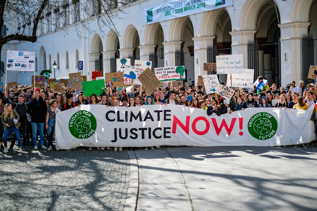 Menschenmenge in einer Stadt bei Sonnenschein, sie tragen ein großes Transparent, auf dem steht: Climate Justice now!
