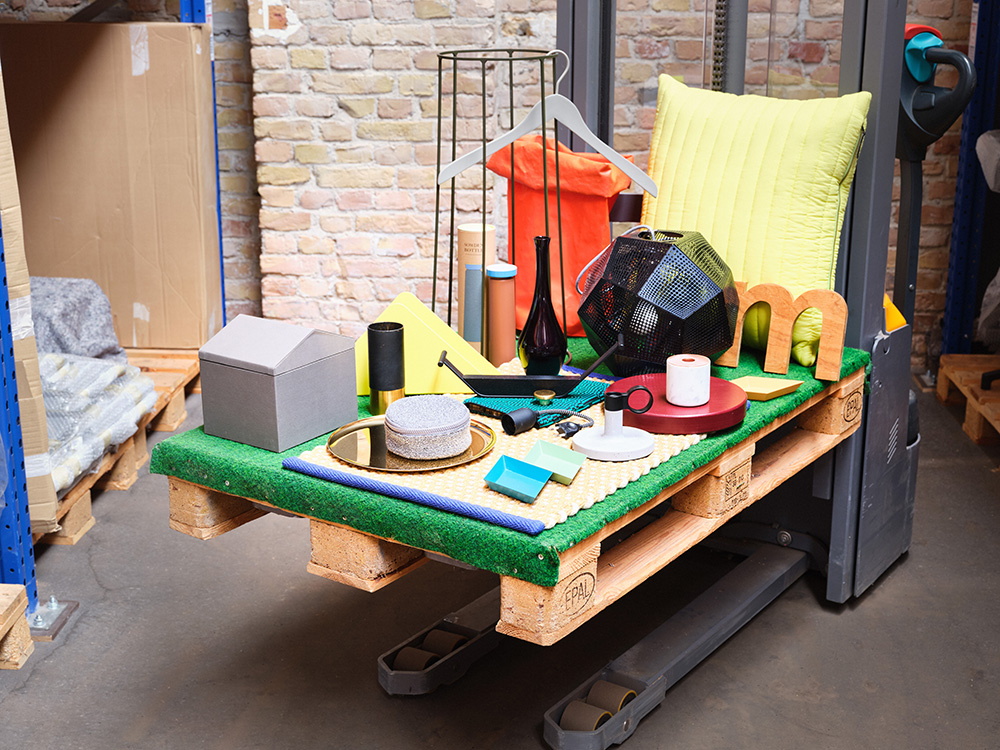 Palette mit einem Stück grünen Kunstrasen auf einem Hubwagen, darauf kleine Design Objekte drapiert wie Kerzenständer, Boxen, Schalen