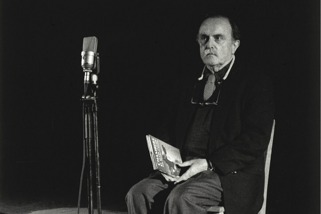 sehr dunkles Schwarz-Weiß-Foto, ein älterer Herr mit Schnurrbart und dunkler Strickjacke sitzt rechts auf einem Stuhl, schaut in die Kamera und hält ein Buch in der Hand, links ein Stand-Mikrofon