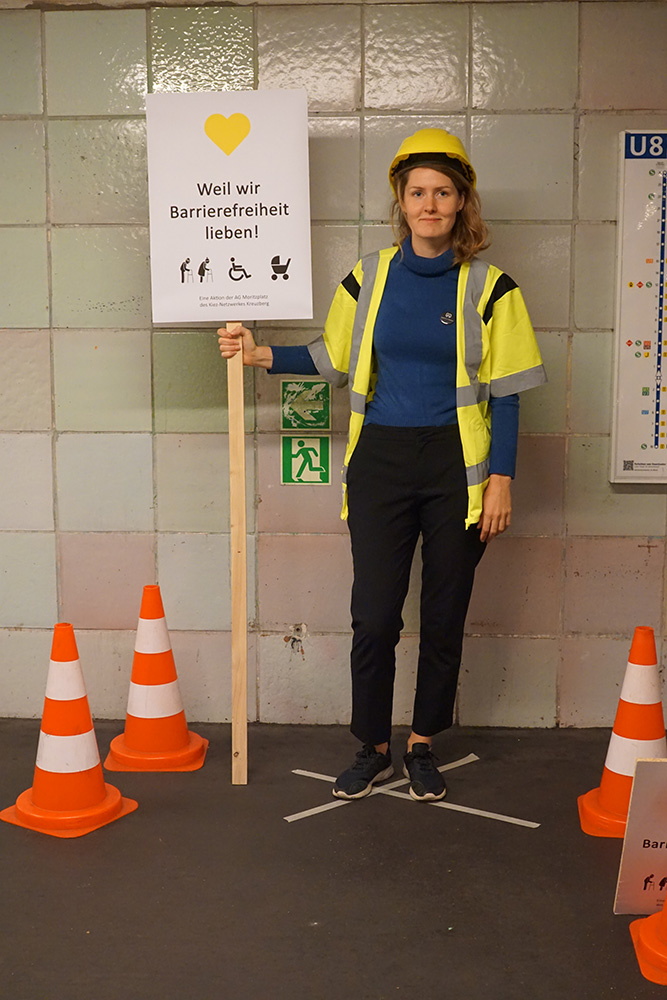 junge Frau mit gelber Warnweste und gelbem Bauhelm hält ein Schild, das an einer Latte befestigt ist, auf dem steht: Weil wir Barrierefreiheit lieben