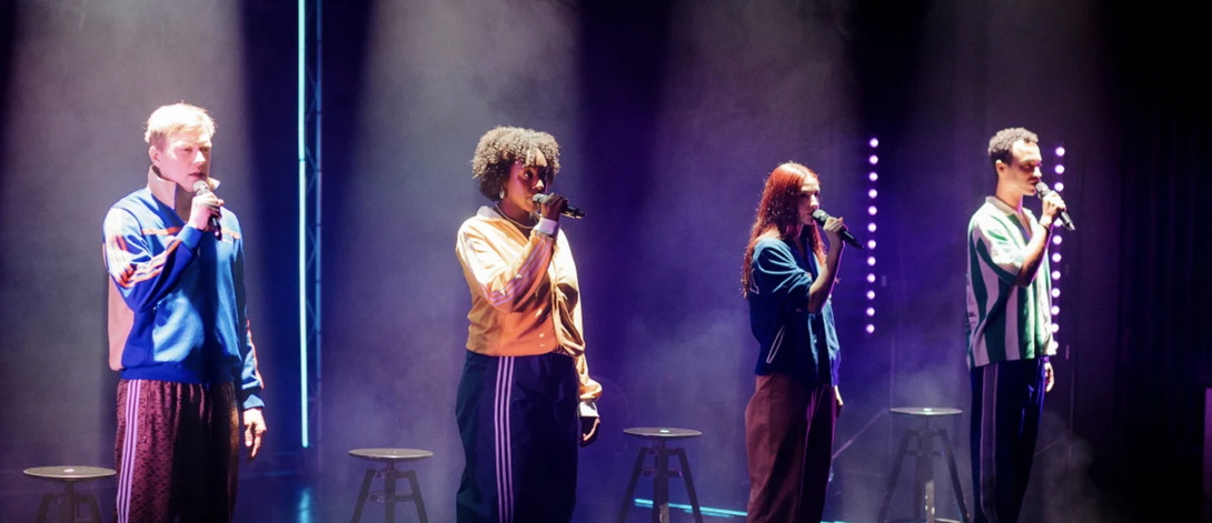 vier junge Menschen in lässiger Kleidung auf Bühne, blau-lila ausgeleuchtet, jede*r einzelne an einem Standmikrofon, stehen in einer Reihe, wahrscheinlich zum Publikum gewandt