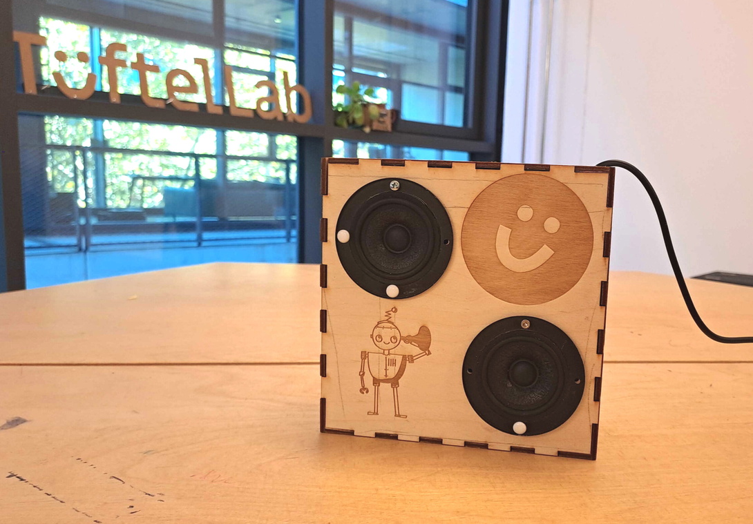 schwarze, runde Lautsprecher in einem Holzgestell, auf dem ein Smiley und ein Roboter zu sehen sind, stehen auf einem Holztisch vor einem großen Fenster