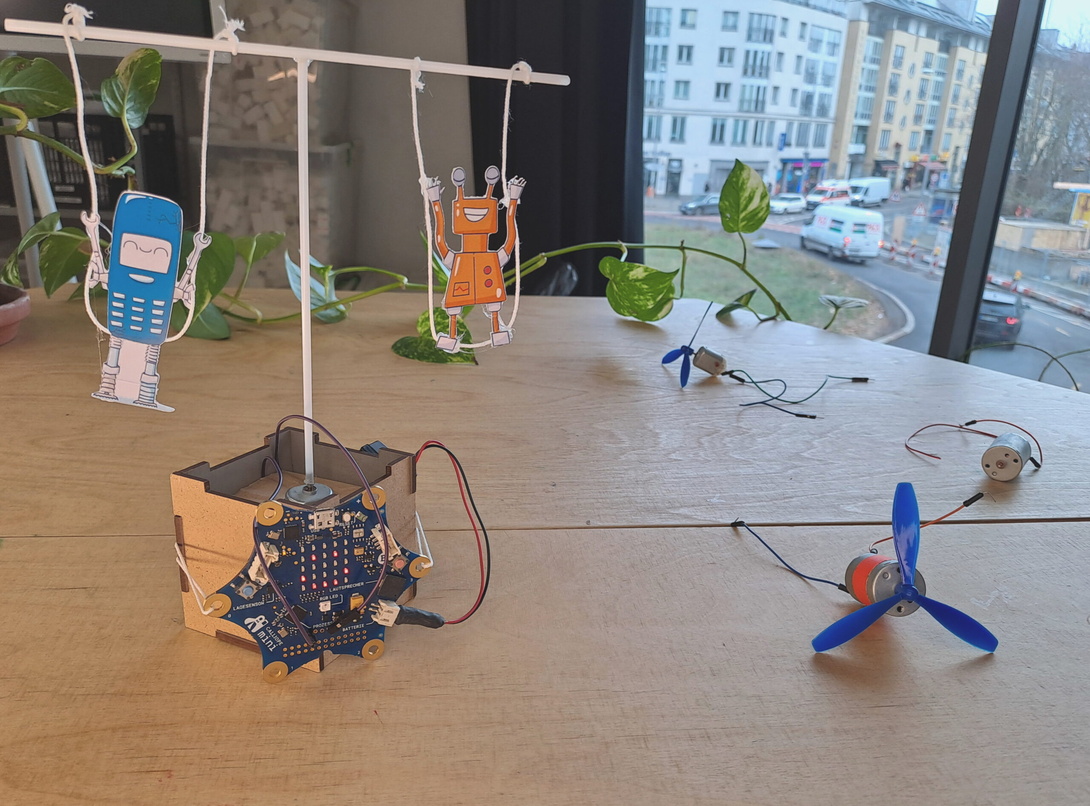 kleine, verkabelte Box mit Holzwänden auf einem Holztisch, darin steckt eine Konstruktion aus einem senkrechten und einem waagerechten Stab, an dem zwei Pappfiguren baumeln wie ein Mobile
