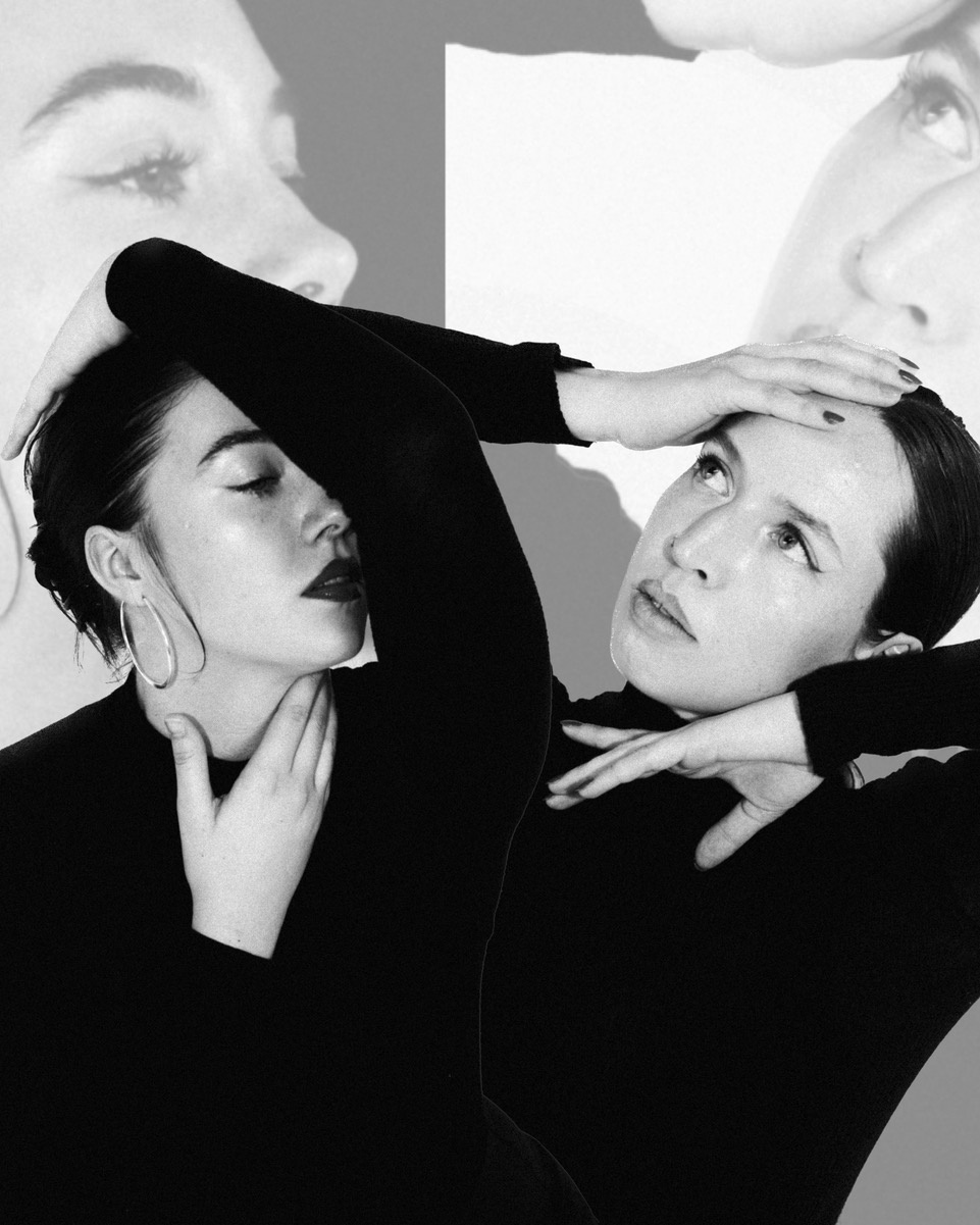 zwei zierliche, weiblich gelesene Personen mit zurückgebundenen dunklen Haaren und schwarzen engen Pullovern in einer engen Bewegung, eine hat die rechte Hand am Kopf, in ihren abgewinkelten Arm schmiegt sich die andere