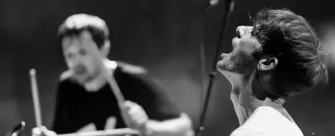 schwarz weiß Foto von zwei Musikern, im Hintergrund ein Mann im schwarzen T Shirt mit Schlagzeugstöcken in Bewegung, vorne ein dunkelhaariger Mann im weißen Shirt, der seinen Kopf in den Nacken legt und den Mund geöffnet hat wie beim Singen oder Schreien