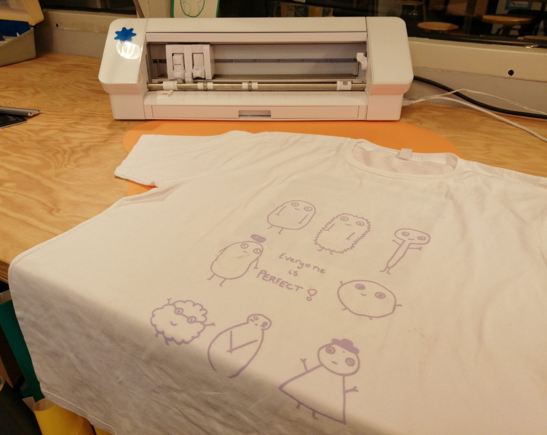 ein weißes T-Shirt liegt auf der Kante eines hölzernen Tisches, bedruckt mit vielen kleinen Figuren, im Hintergrund ein weißes Gerät