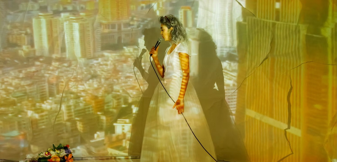 gelblich ausgeleuchtetes Bild, eine Frau in einem weißen Hochzeitskleid vor einer Videoprojektion eines urbanen Settings