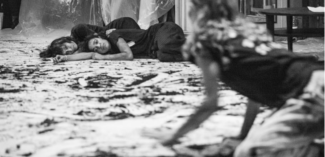 schwarz weiß Foto, zwei Frauen liegen in dunklen Shirts auf einem Boden, der mit Stoff oder Papier ausgelegt zu sein scheint, eine andere Frau sehen wir von hinten, die krabbelt in Richtung der beiden 