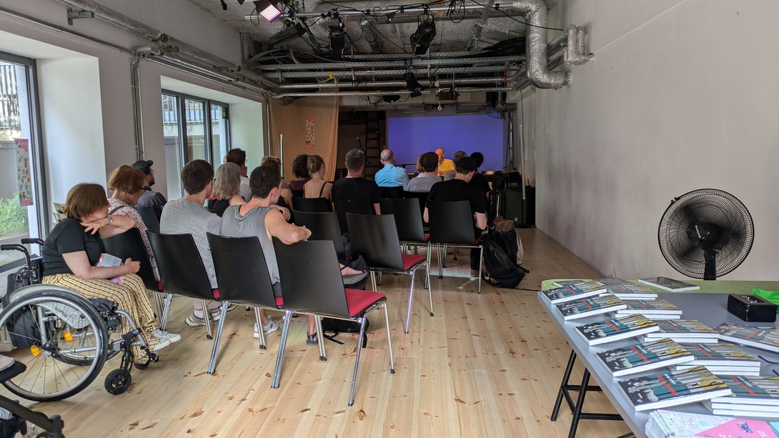 Publikum von hinten in einem Raum mit hellem Parkett und links einer Fensterfront, vorne ein Mensch im Rollstuhl