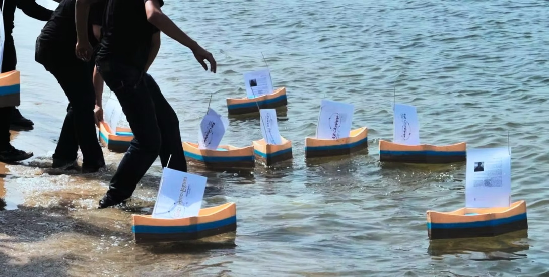 kleine gebastelte Schiffchen mit Papiersegeln werden von einigen Menschen in schwarzen Anzügen, deren Köpfe im Bild nicht zu sehen sind, ins Wasser gelassen
