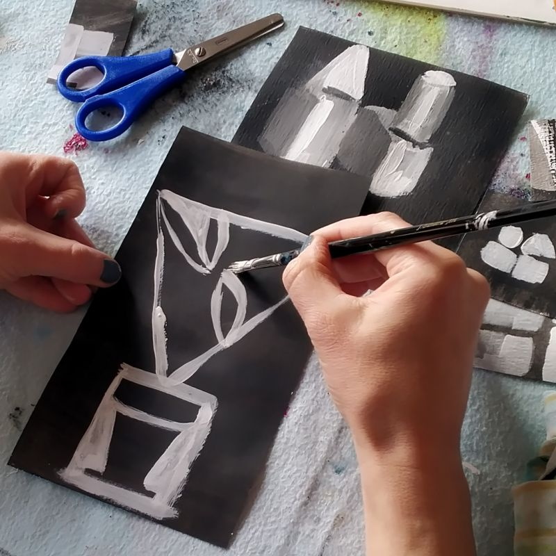 schwarzes Tonpapier, eine weiße Hand malt mit Pinsel und weißer Farbe Formen darauf