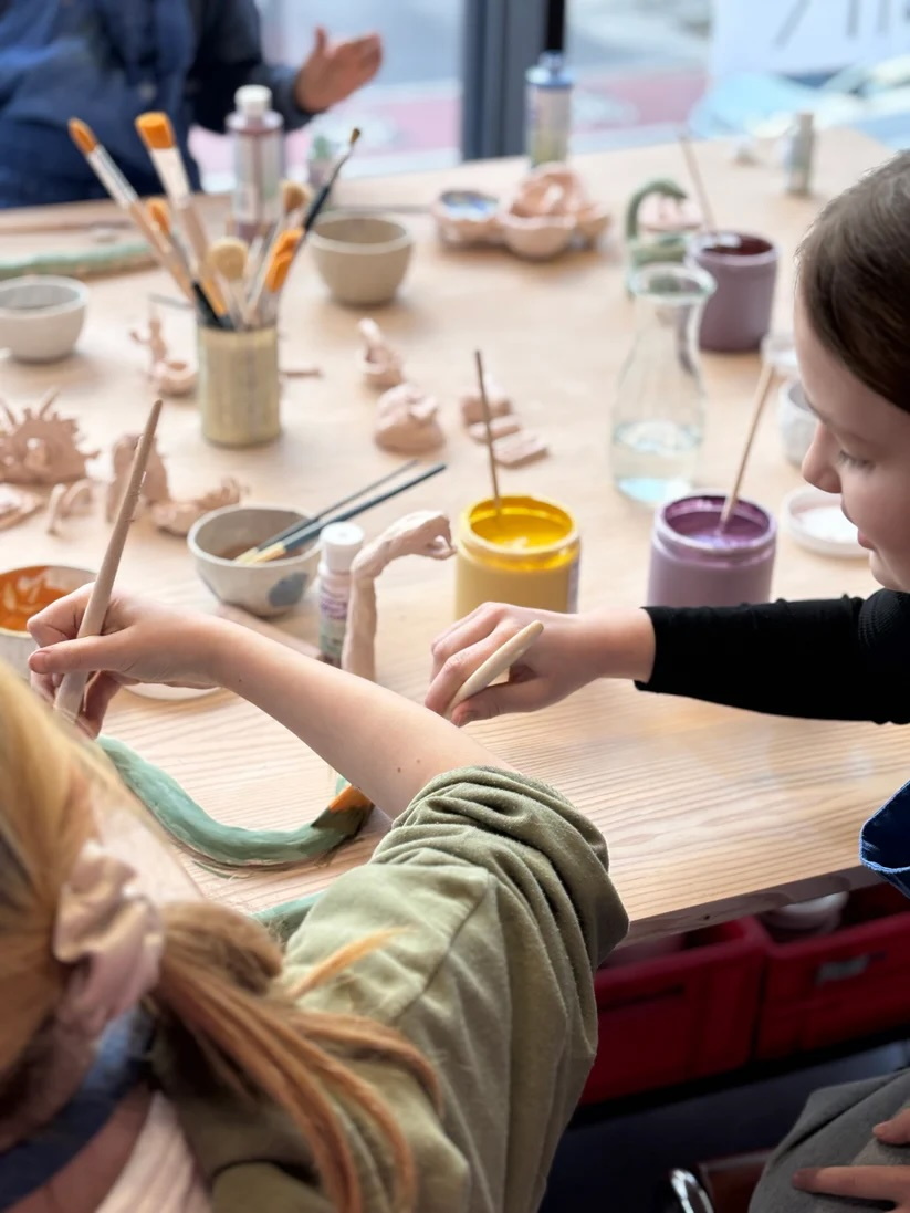 Holztisch vollgestellt mit Arbeitsmaterialien, Pinsel, Farbe, Keramik Objekte, zwei Mädchen im Anschnitt, die Keramik bemalen