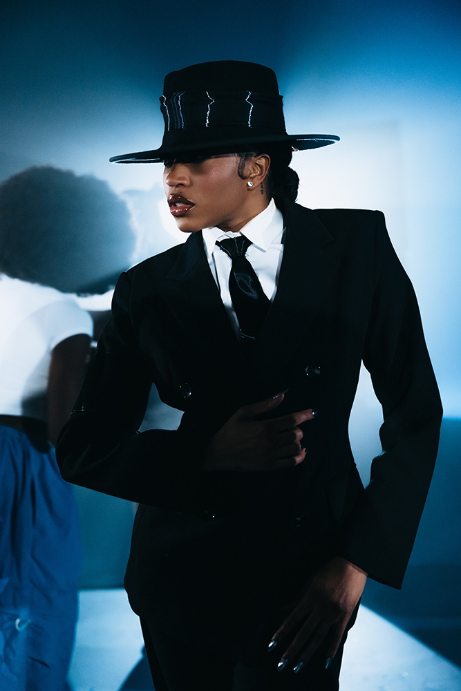 schwarze Person mit tief in die Stirn gezogenem Hut im schwarzen Anzug mit weißem Hemd, schwarze Krawatte, Jackett zugeknöpft, rechte Hand in die Hüfte gestemmt, die linke in der Hosentasche, herausfordernde Pose