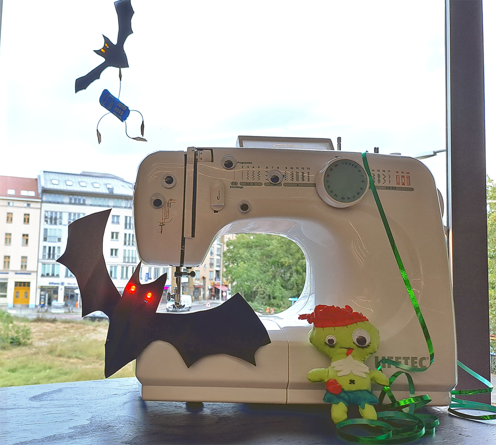 kleine weiße Nähmaschine steht vor einem Fenster, geschmückt mit einer gebastelten Fledermaus mit leuchtenden Augen und einer kleinen grünen Puppe
