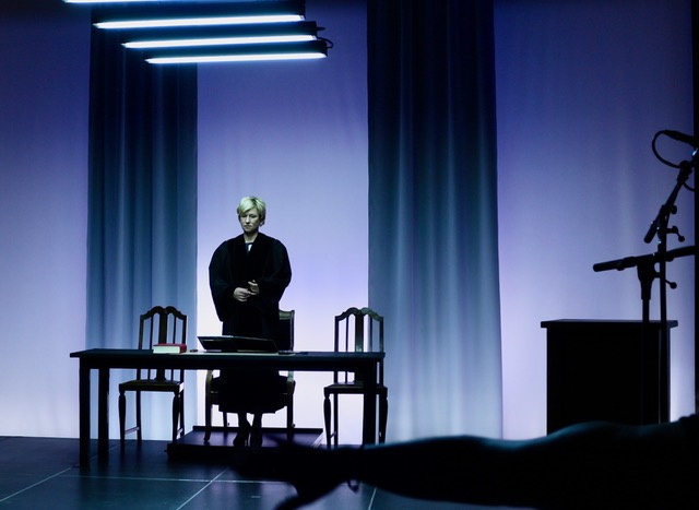 mittelalte blonde Frau im Richterinnen-Talar steht hinter einem Tisch in einem blau ausgeleuchteten Raum