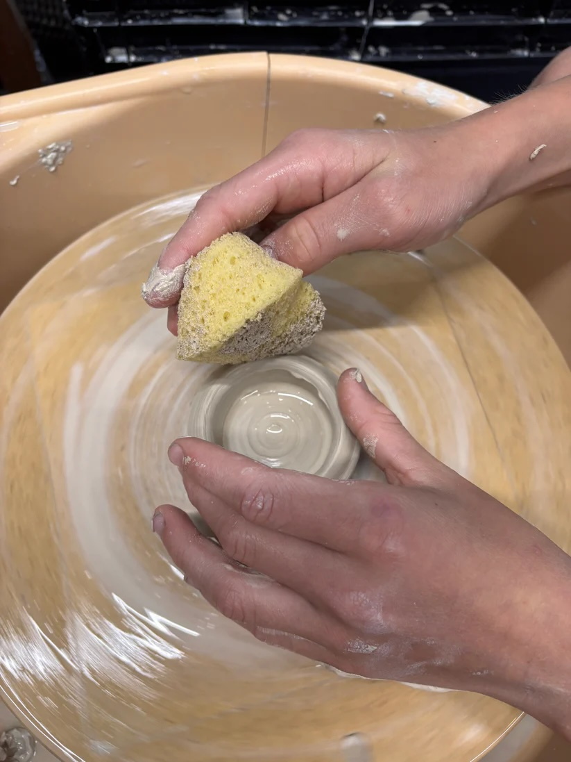 zwei Hände bearbeiten feuchten Ton in Kreisform, der sich dreht, mit einem kleinen gelben Schwamm