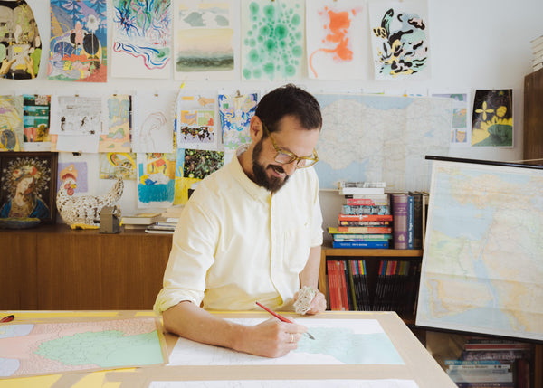 mittelalter weißer Mann mit langen dunklen Haaren und Vollbart, steht am Tisch und malt mit einem Pinsel, im Hintergrund Bilder an der Wand