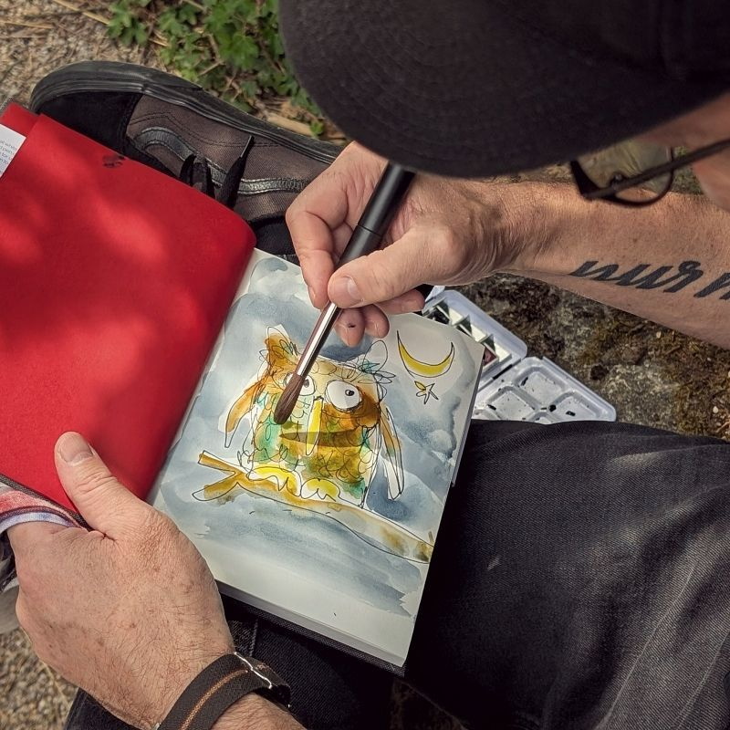 eine Person mit dunkler Schiebermütze und dunkler Brille seitlich im Anschnitt, Betrachter schaut auf die Skizze einer Eule, die gelb aquarelliert wird