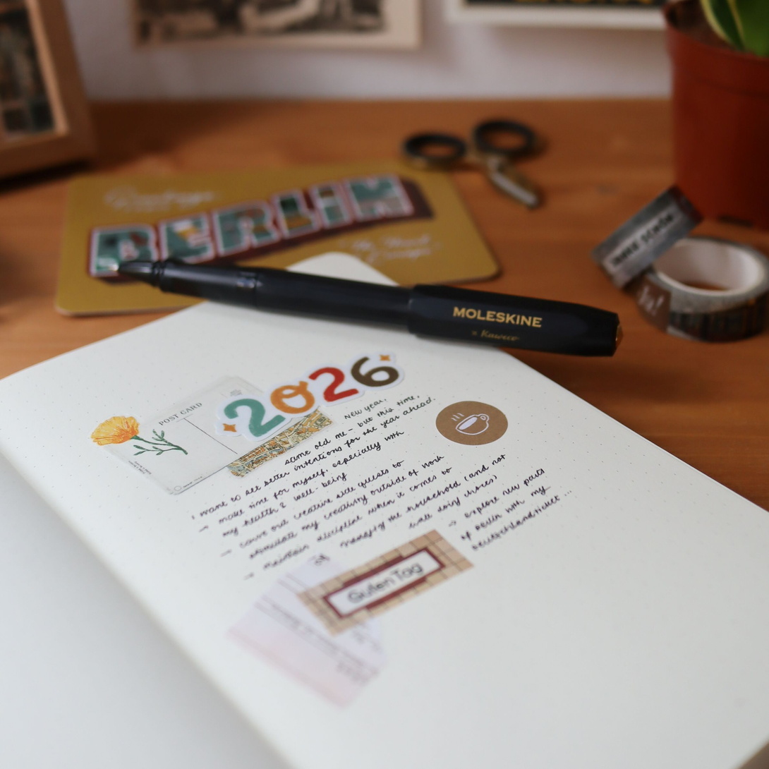 aufgeschlagenes Notizbuch mit einem ersten Eintrag mit einer bunten 2026 und einem schwarzen Moleskine Stift, der auf dem Papier liegt