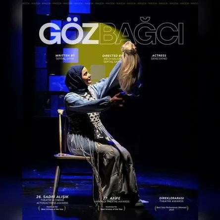 junge Frau mit Kopftuch und langem Gewand sitzt auf einem Stuhl auf einer Bühne, sie hält eine langhaarige Perücke vor sich in die Höhe