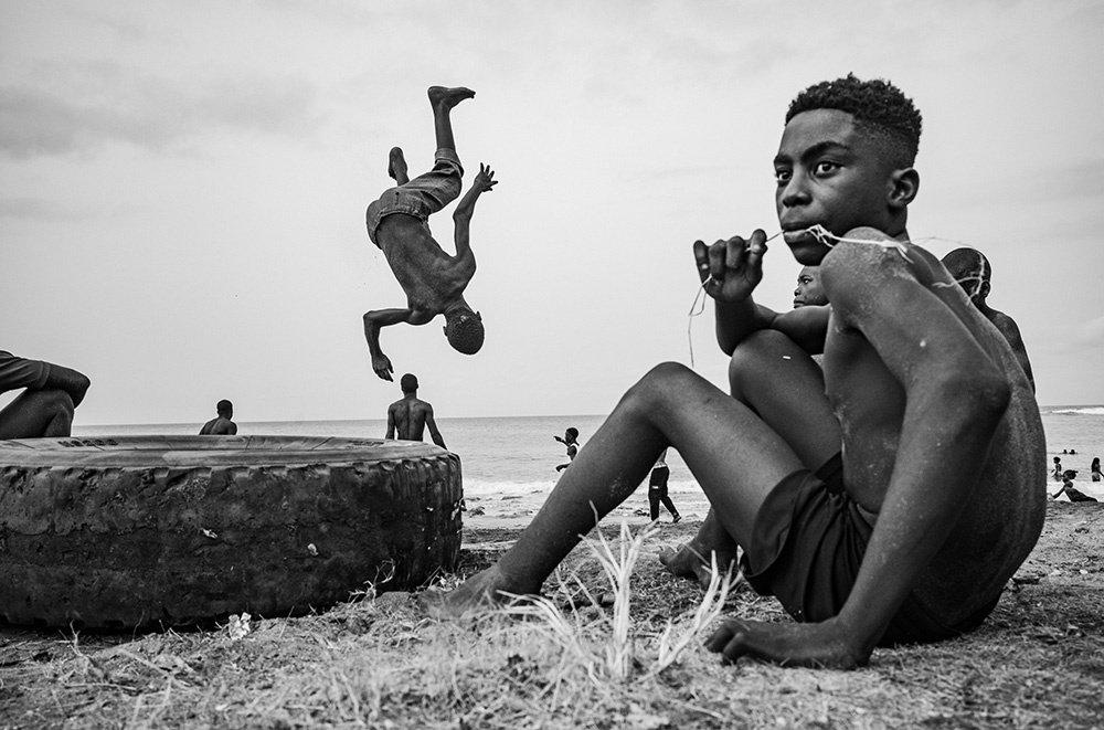 schwarz weiß Foto, im Vordergrund ein Jugendlicher mit freiem Oberkörper neben einem Autoreifen, im Hintergrund ein Junge in einer Salto Bewegung in der Luft, dahinter das Meer mit einigen Menschen