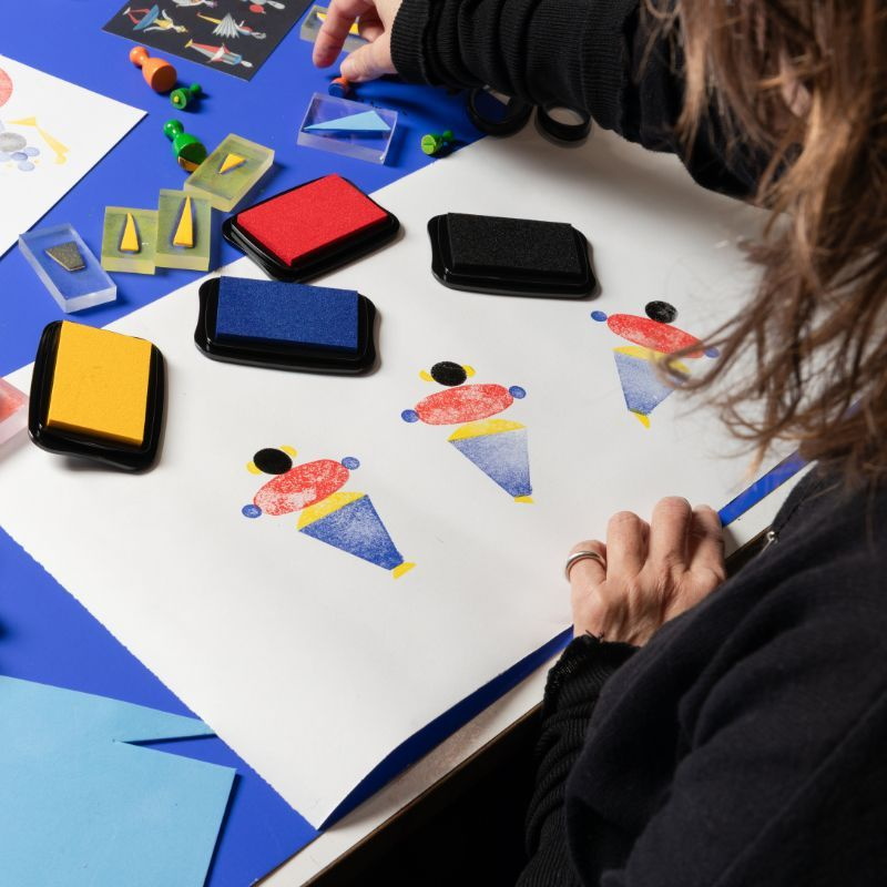 vier Stempelkissen gelb, blau, rot und schwarz auf einem großen weißen Papier, Dreiecke und Ovale gestempelt, so dass sich Figuren ergeben 