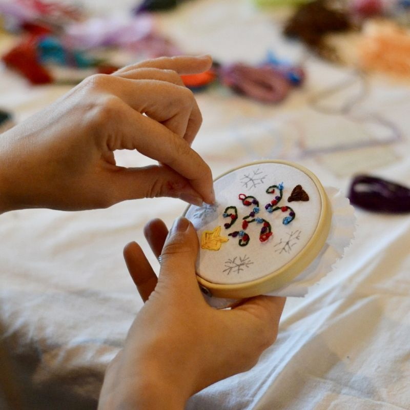 zwei Hände besticken eine kleine kreisrunde Form, die mit hellem Stoff bezogen ist, florales Motiv