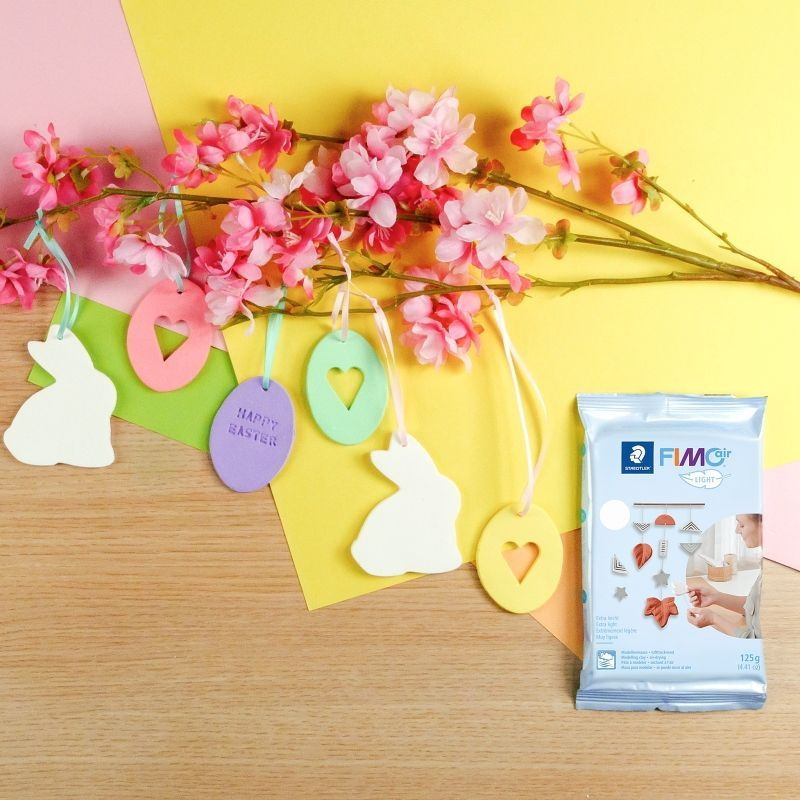 gelbes Papier, darauf liegt ein Ast mit Blüten, daran Oster-anhänger, zwei weiße Hasen, drei Eier in verschiedenen Farben