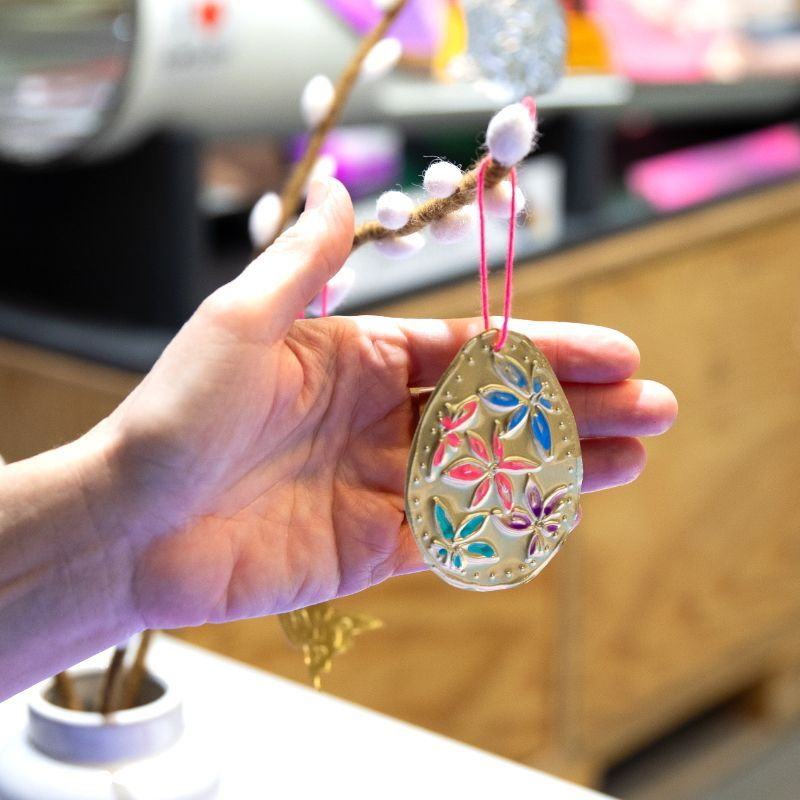 eine Hand greift hinter einen eiförmigen, geprägten Anhänger mit bunten Blumen, der an einem Ast hängt