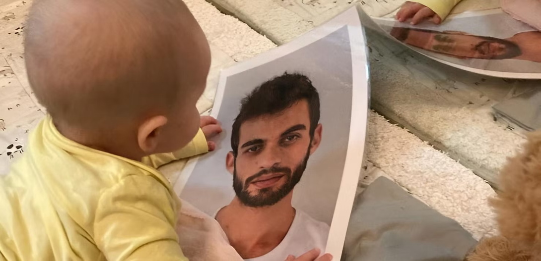 ein kleines haarloses Baby in gelbem Body auf dem Boden, von hinten fotografiert, in den Händen hält es das große Bild eines jungen Mannes mit kurzen dunklen Haaren und einem dunklen Bart in weißem Shirt
