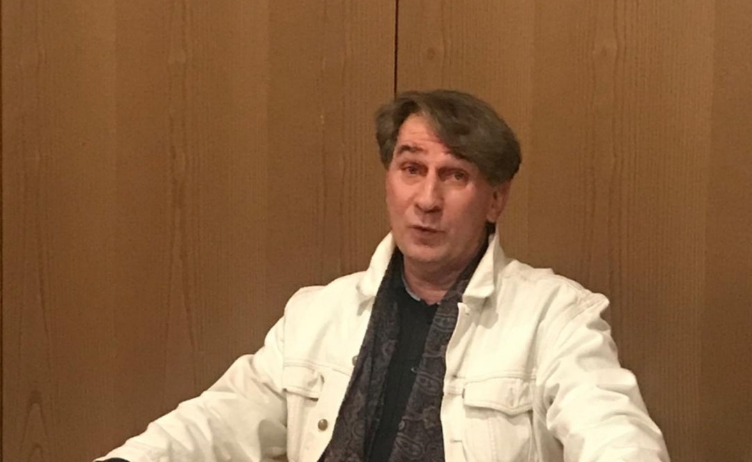 mittelalter weißer Mann mit dunklen, mittellangen Haaren in einer weißen Jeansjacke, darunter ein dunkles Shirt, sitzt vor einer Holzwand