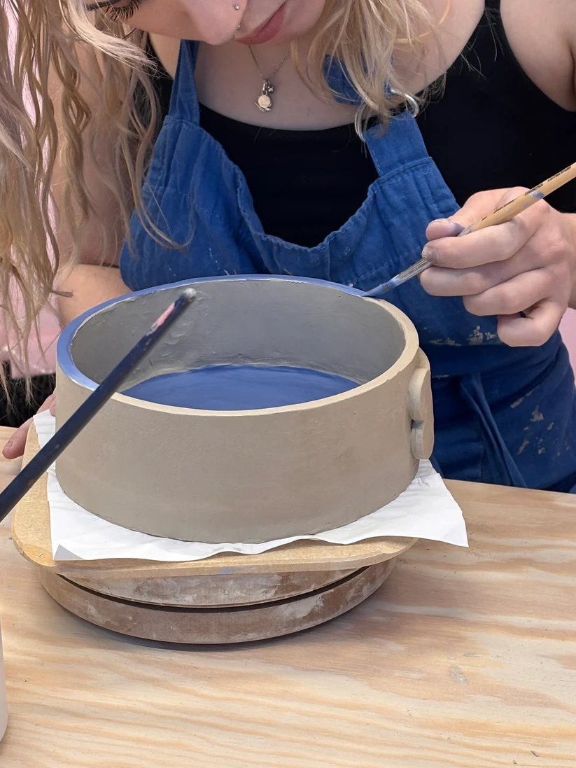 eine junge blonde Frau mit blauer Latzhose und langen welligen Haaren beugt sich nach vorne über einen Holztisch, auf dem eine Schale aus Naturton steht, die sie gerade mit einem Pinsel bemalt