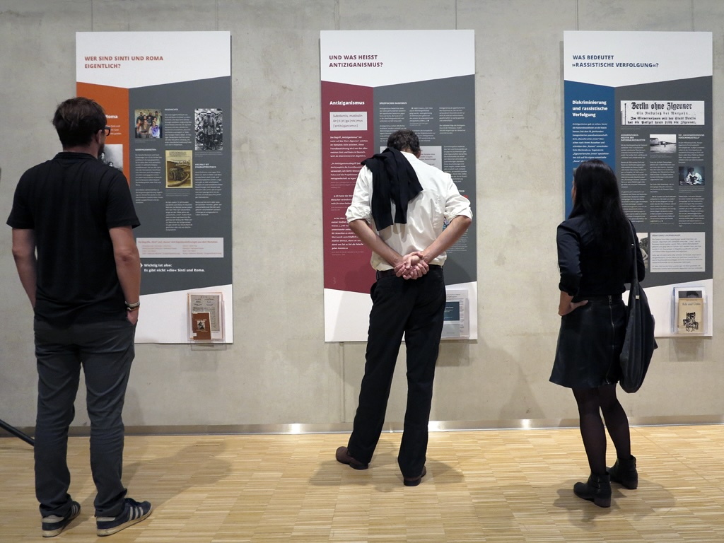 drei Menschen von hinten fotografiert, die großformatige Ausstellungsposter betrachten und lesen