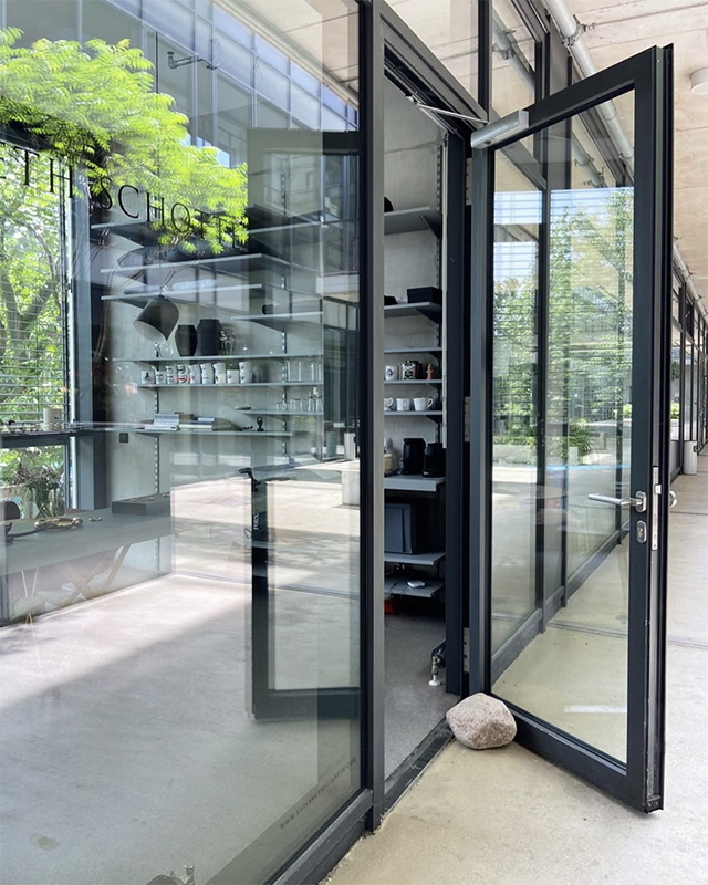 gläserner Raum mit leicht geöffneter Glastür, diese mit dunkelgrauer Rahmung, man schaut auf ein Regal und erkennt gegenüber der Tür ein anderes bodentiefes Fenster