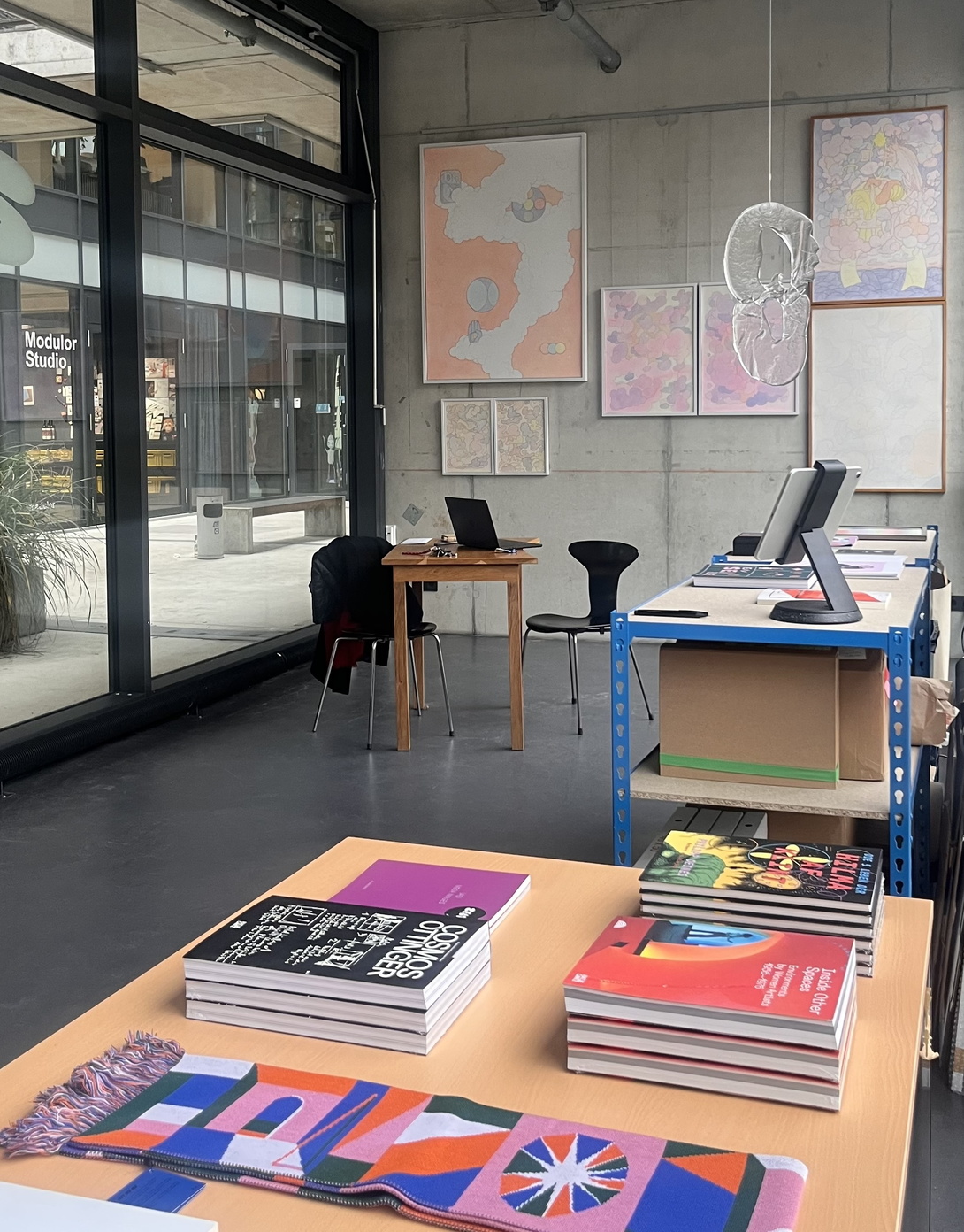 heller Raum mit einer Glasfront, ein Büchertisch im Vordergrund, hinten verschieden große Bilder an einer Betonwand