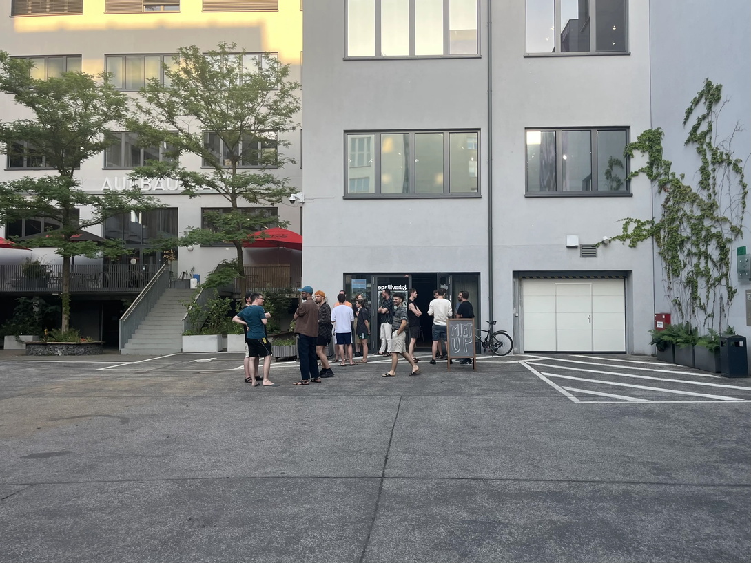 eine Hoffläche vor einem hellen Neubau, eine Gruppe Menschen im Gespräch, im Hintergrund Bäume